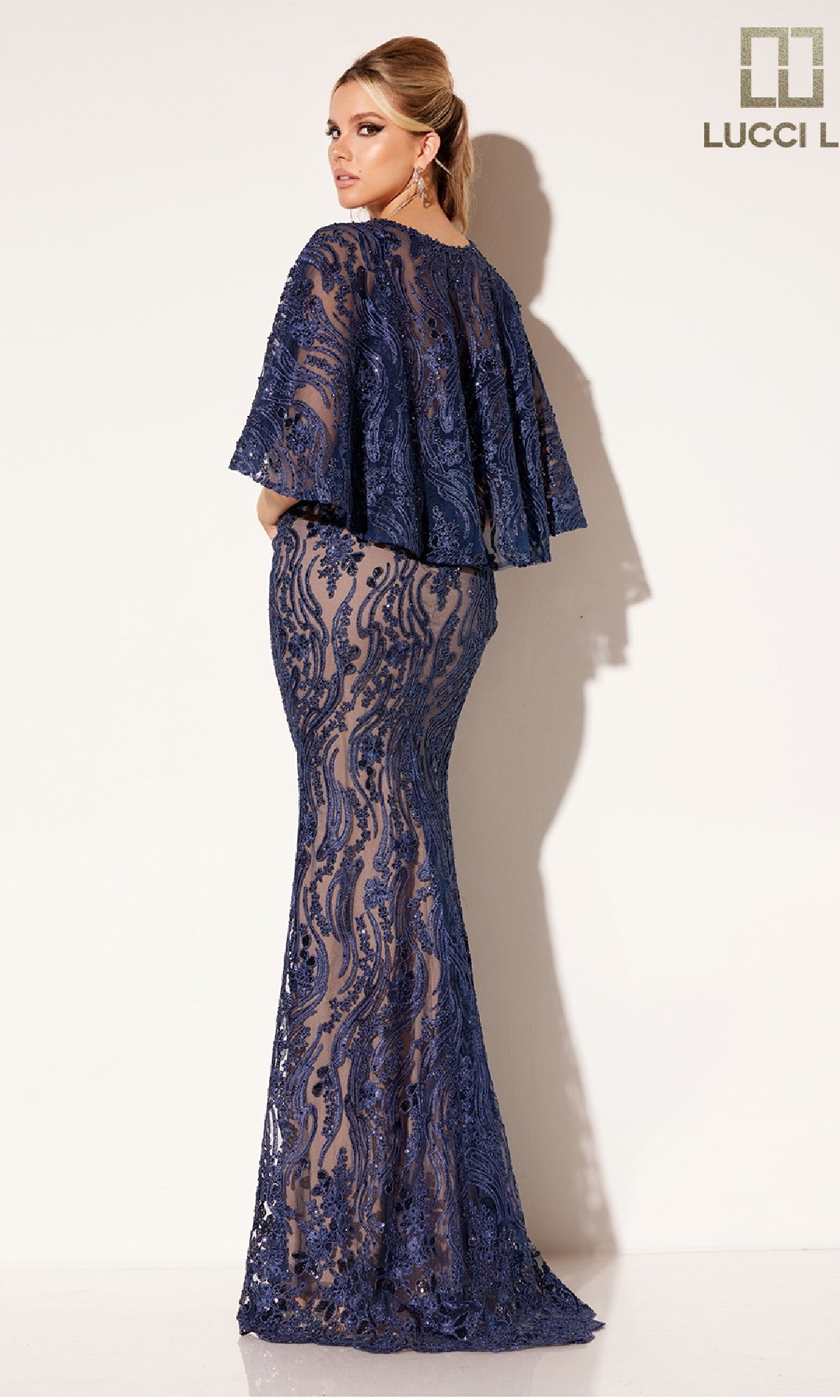 Woman wearing the Lucci Lu C8086, a fitted navy blue formal evening gown featuring lace, a sheer overlay, and an elegant capelet, stands against a plain light background.